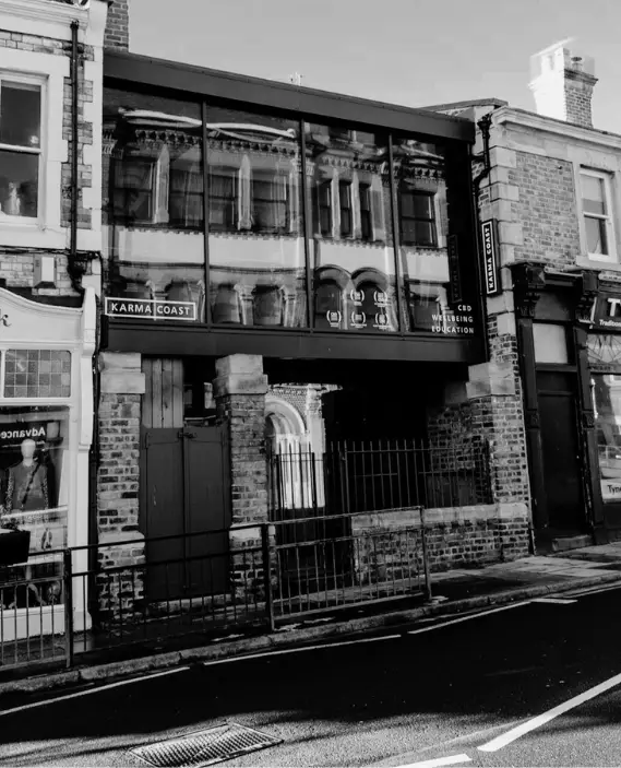 A black and white photo of a brick building with large glass windows reflecting the ornate façade of another building across the street. Signs on the building read Karma Coast and CBD Wellness Location.