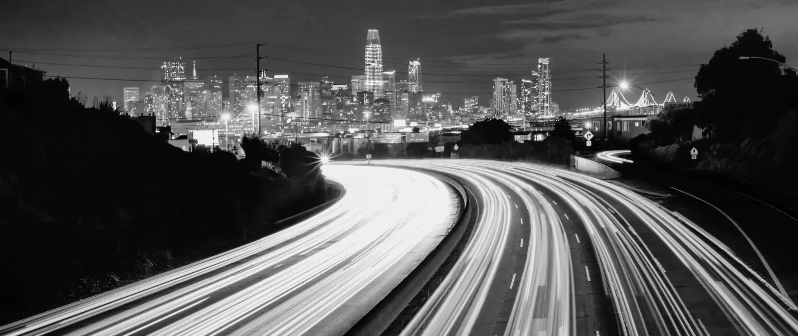 Black and white photo of a city skyline at night with tall buildings in the background and blurred light trails of cars on a curving, multi-lane motorway in the foreground.