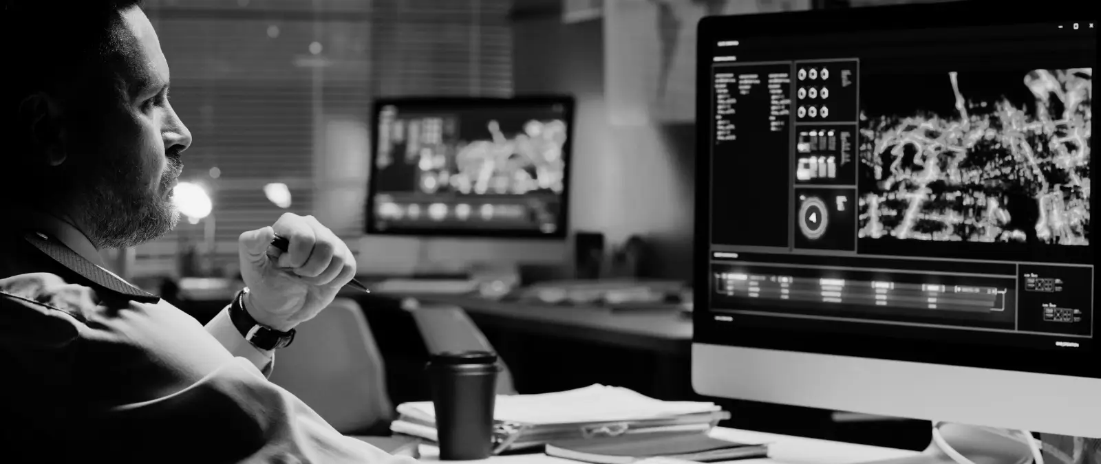 A person sits at a desk in a dimly lit office, studying medical images on a computer screen. Another monitor with charts and documents—perhaps illustrating how topic clusters boost SEO—is visible in the background. A coffee cup and papers are on the desk.