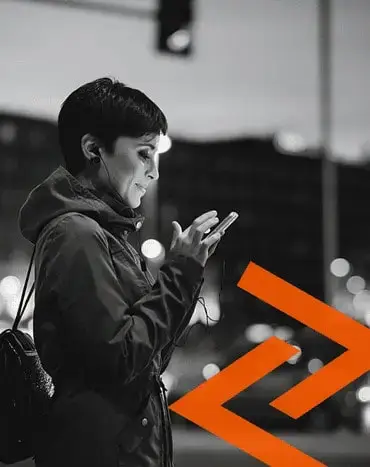 A person with short hair, wearing headphones and a jacket, uses a mobile phone outdoors at dusk. An orange double-arrow graphic is overlaid at the bottom right.