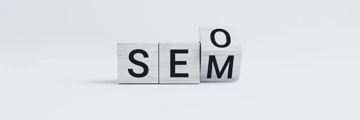Three wooden blocks with the letters S, E, and M are arranged side by side. A fourth block with the letter O is resting on top of the M block, forming the word SEO vertically and SEM horizontally.