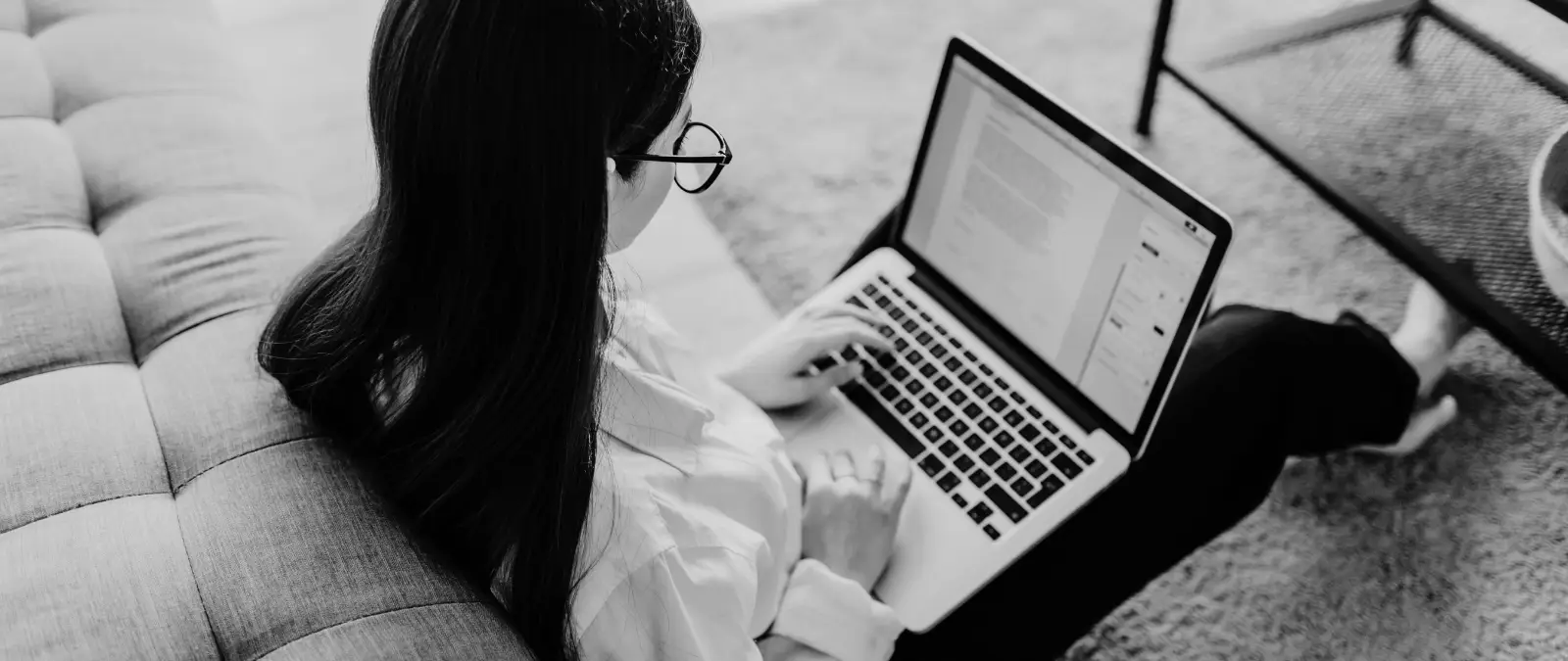 A person with long hair and glasses sits on the floor next to a sofa, typing on a laptop with documents open on the screen. The setting is cosy with a soft carpet and a nearby coffee table.
