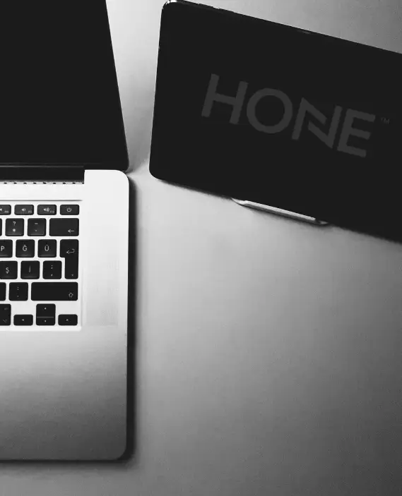 Black and white image showing part of a laptop keyboard on the left and a device displaying the word HONE on the right, both on a flat surface.