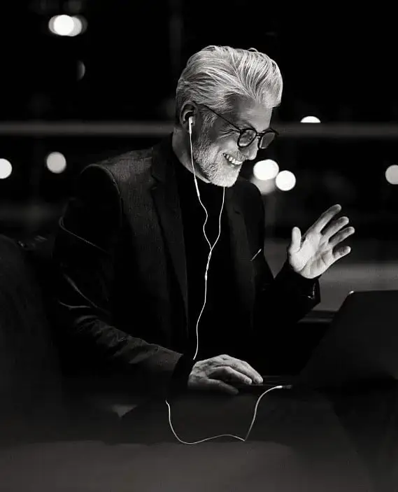 A smiling older man with glasses and earphones waves at a laptop screen during a video call, sitting in a dark, modern setting.