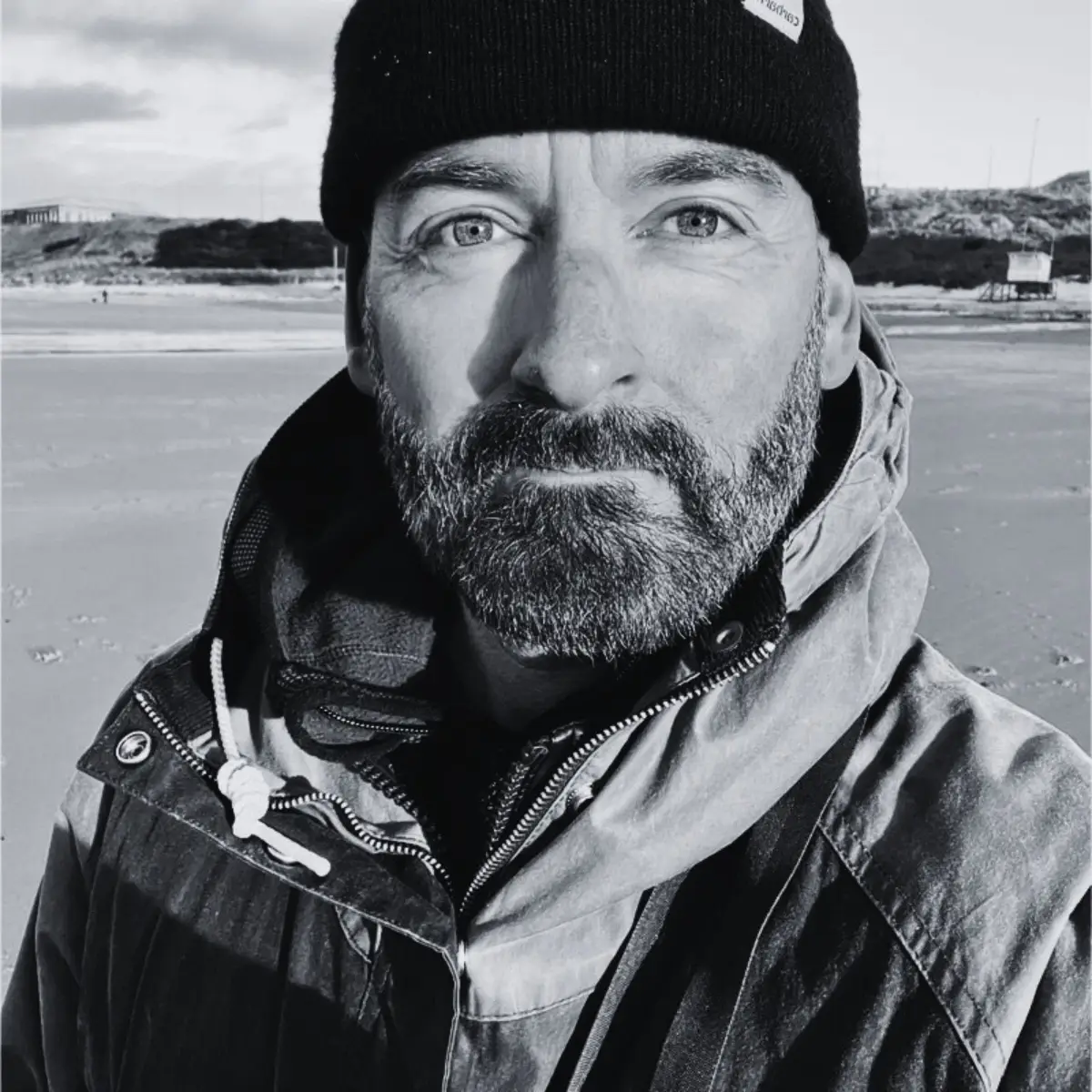A bearded man in a jacket and beanie stands on a beach, looking at the camera. The image is in black and white, with sand and a blurred shoreline visible in the background.