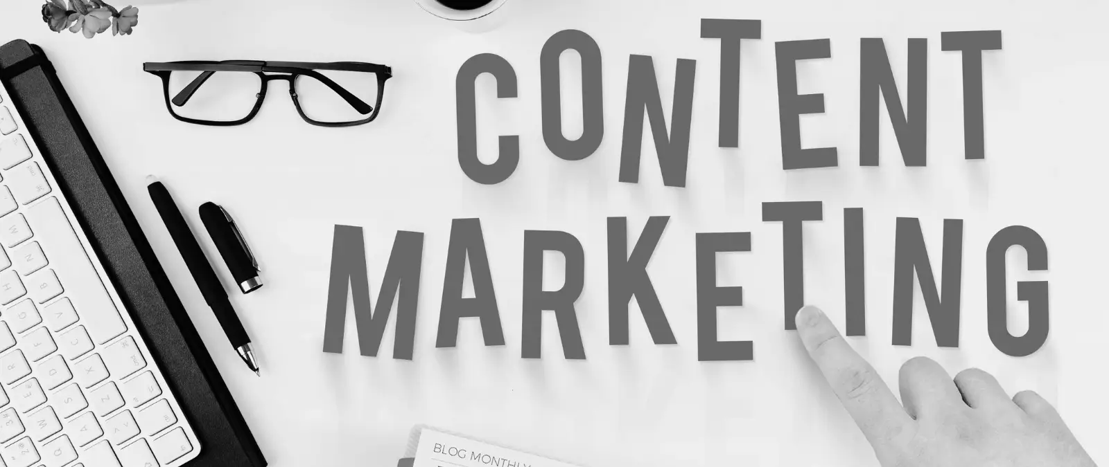 Black and white image showing a desk with glasses, pens, keyboard, and a hand pointing at large text reading “CONTENT MARKETING.” Subtle elements suggest the strategic power of content organisation, illustrating how topic clusters boost SEO within a well-structured workspace.