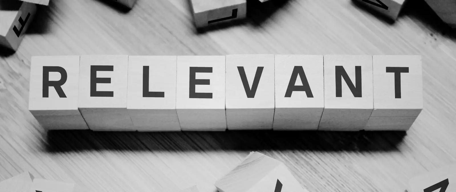 Wooden blocks arranged in a row spell out the word RELEVANT in bold, black letters. The background is a wooden surface, and the image is in black and white.