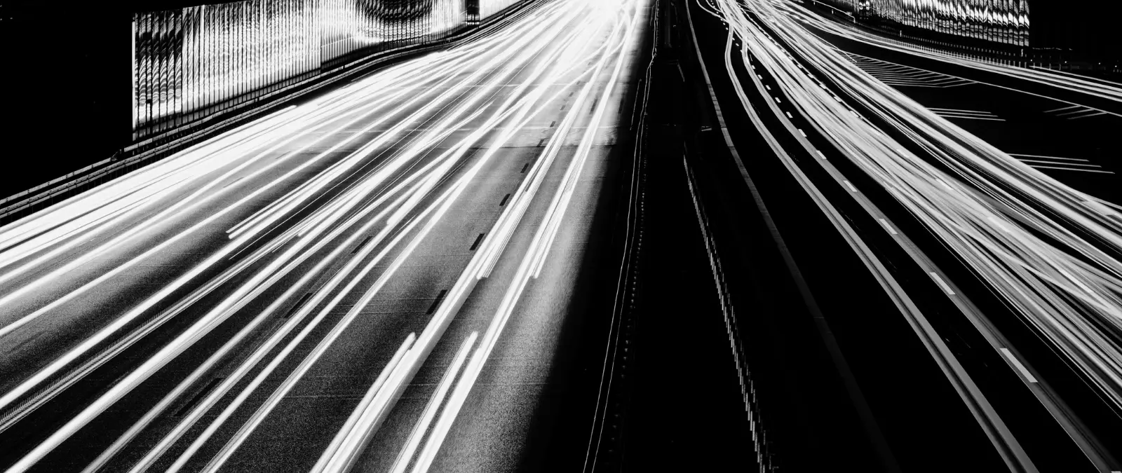 Black and white photo of a motorway at night, showing long-exposure light trails from fast-moving vehicles in both directions, creating bright lines against the dark carriageway.