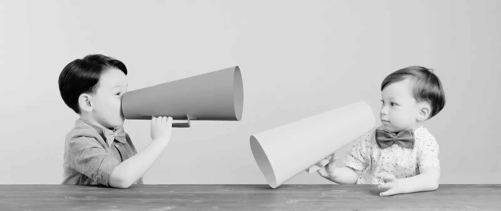 Two young boys sit at a table facing each other, each holding a large paper megaphone to their mouths, as if pretending to shout or talk through them. The image is in black and white.