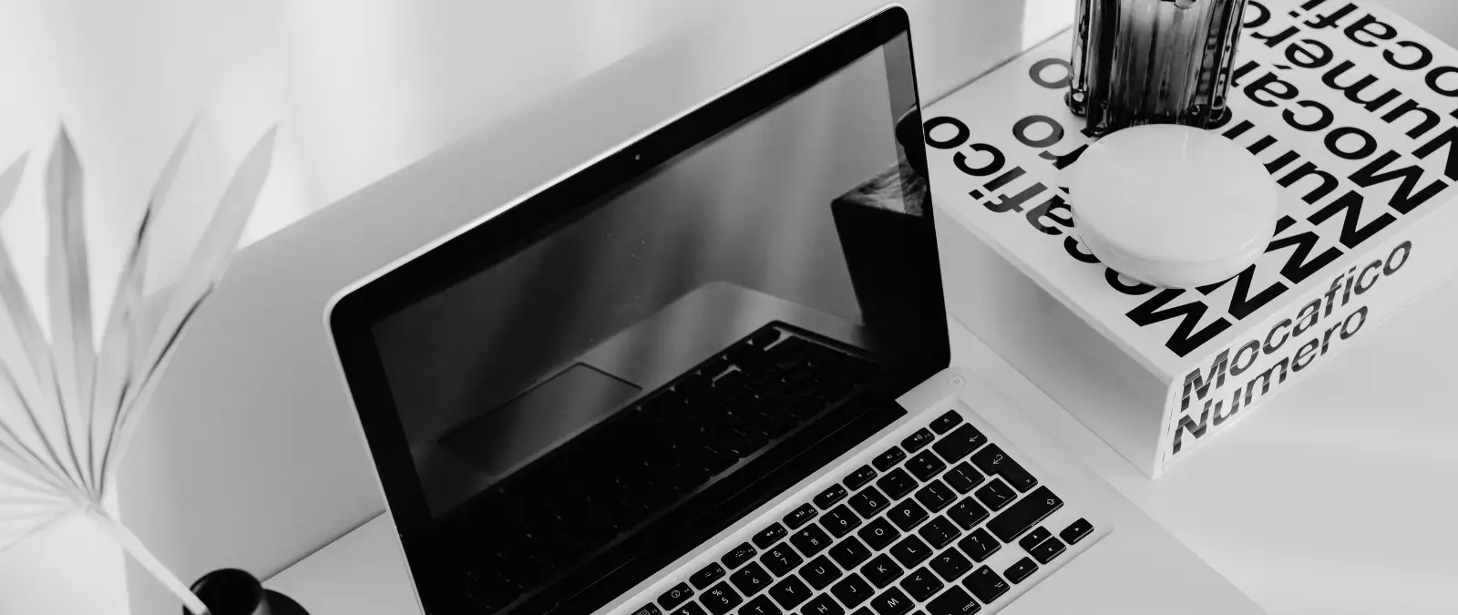 A black and white photo of a laptop on a desk next to a plant, a glass, a white round object, and a book titled Mocafico Numero. The laptop screen is off, reflecting part of the desk setup—an ideal minimalist workspace for focused tasks like on-page SEO optimisation.