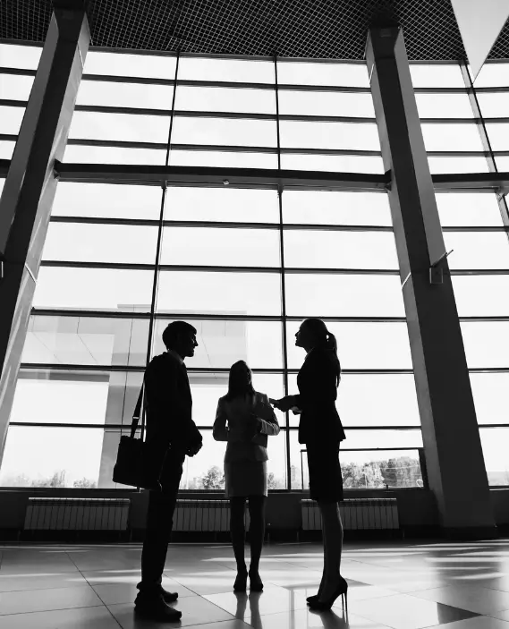 Three people in business attire stand and talk in a modern building with large windows, silhouetted against the bright natural light coming from outside.