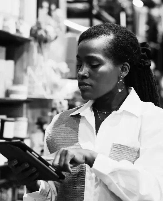 Black and white photo of a woman in a white shirt using a digital tablet in a shop or workspace, surrounded by shelves with various objects.